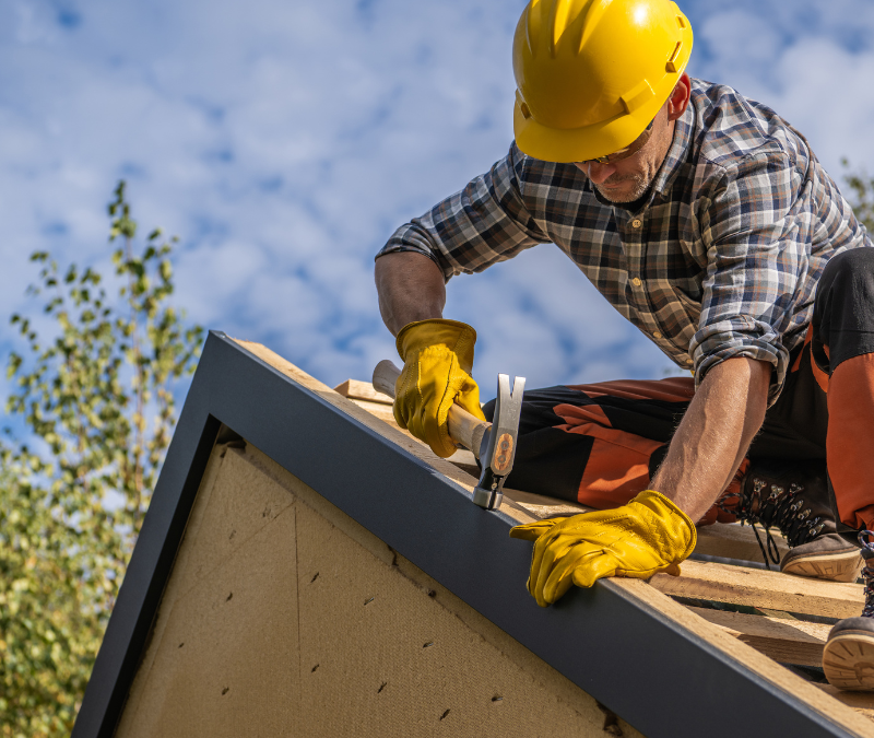 A man wearing a hard hat and yellow gloves is working on a roof.