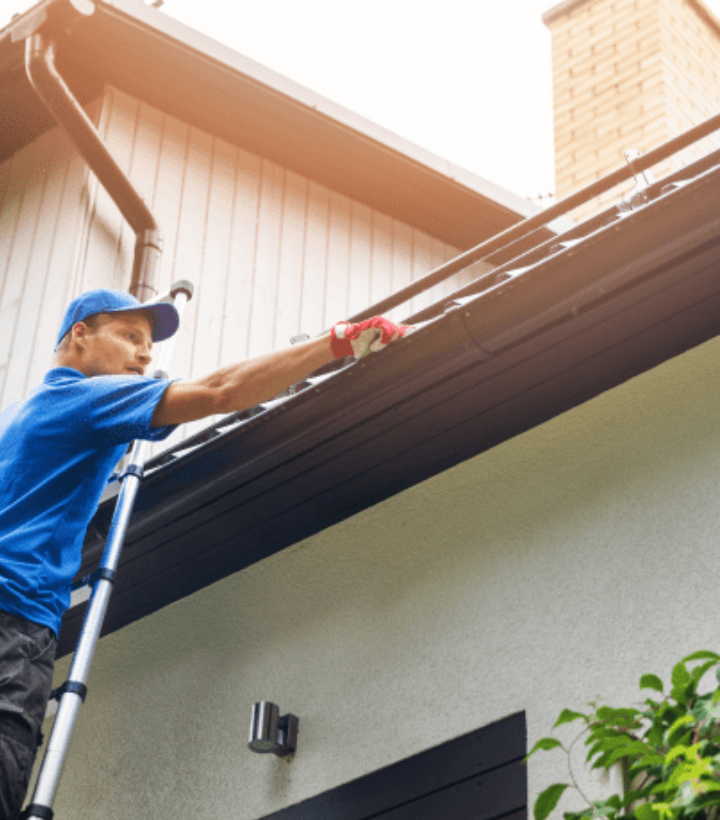A man is standing on a ladder working on the gutters of a house.