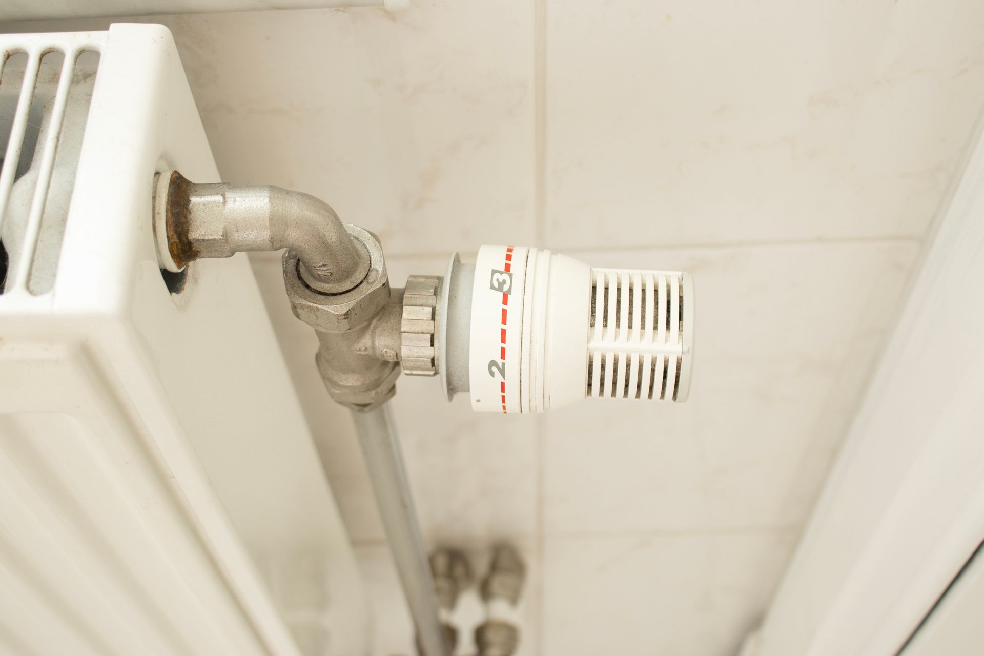 A white radiator with a white thermostatic valve set between numbers 2 and 3, mounted against a white tiled wall.