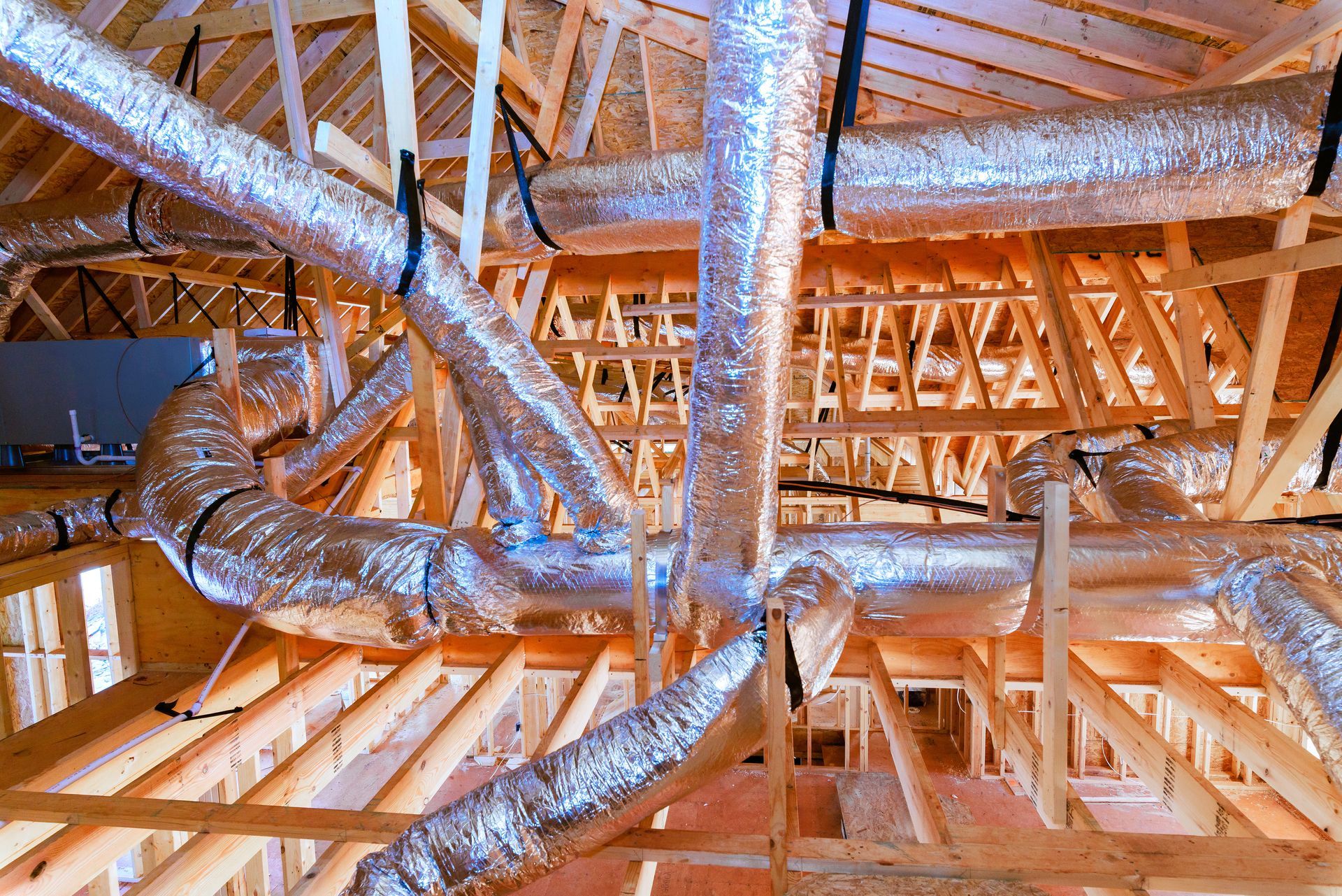 Insulated silver HVAC ducting installed throughout the wooden attic framework of a building under construction.