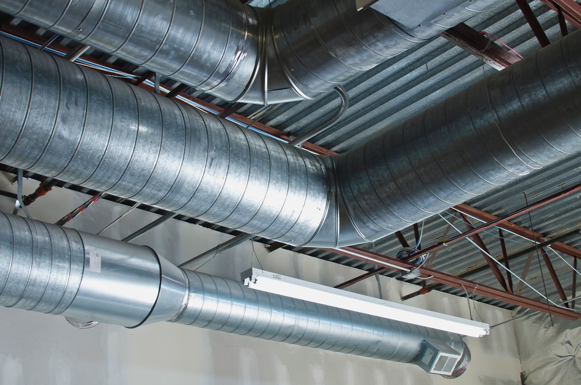Exposed metallic ventilation ducts and a light fixture mounted beneath a corrugated industrial ceiling.