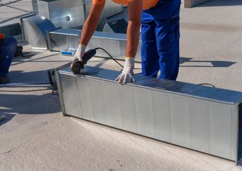 A person in work clothes uses an angle grinder to cut a metal HVAC duct on a construction site.