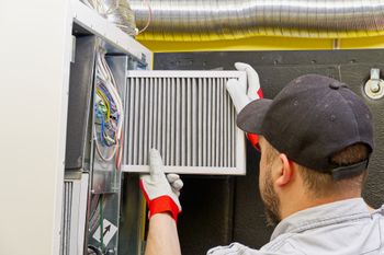 A person wearing gloves inserts a replacement air filter into a furnace unit.