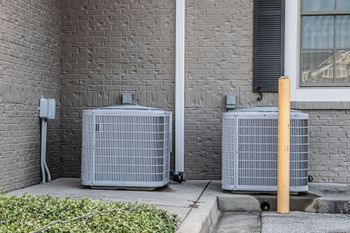 Two gray central air conditioning units sit on a concrete pad against a brick exterior wall next to a yellow bollard.