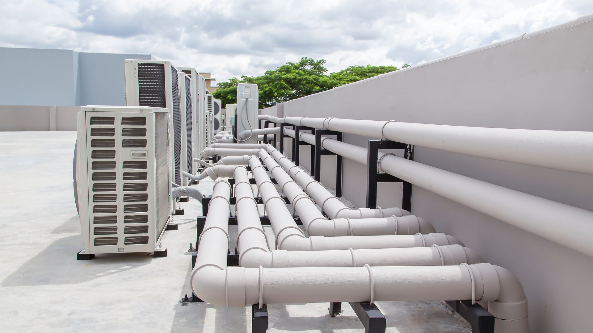 HVAC units and insulated pipes lined along a flat rooftop under a cloudy sky.