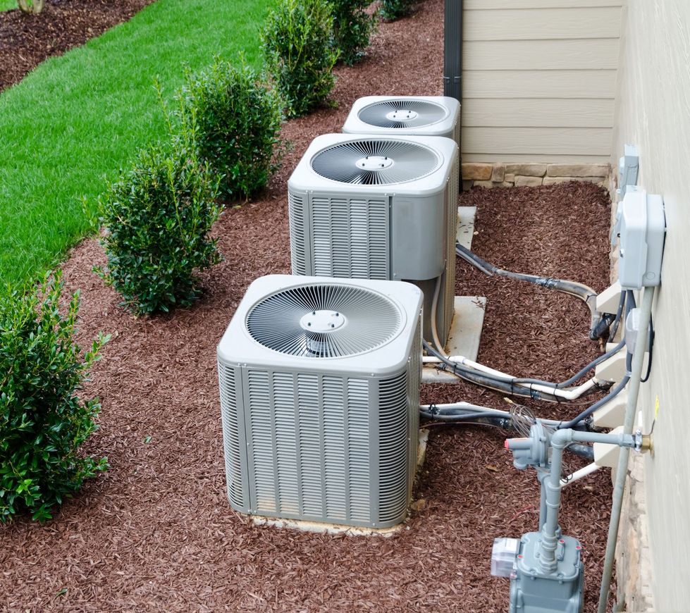 Three air conditioning units arranged in a row on a bed of mulch next to a house wall and a grassy lawn.