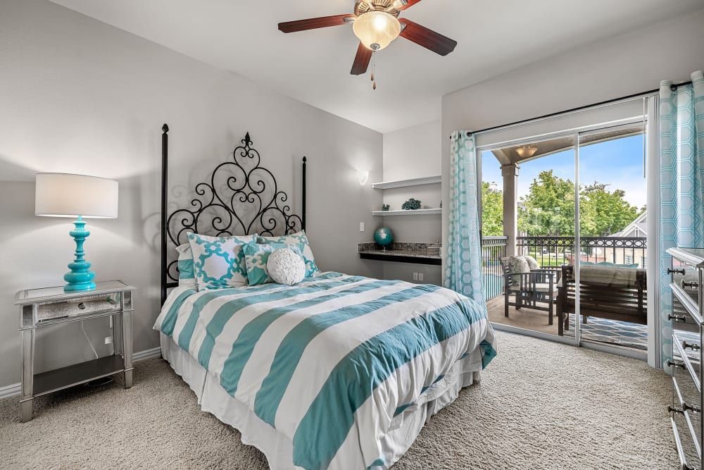 A bedroom with a bed, nightstand, lamp, window, and ceiling fan at Marquis at Silver Oaks in Grapevine, TX.
