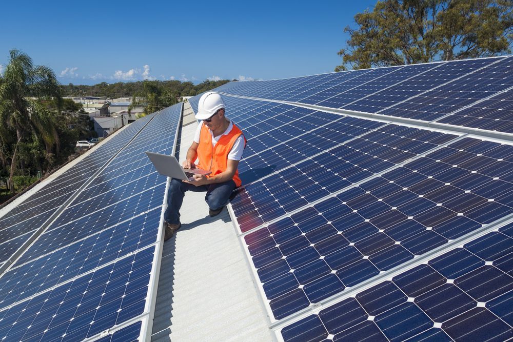 A Man Is Kneeling On Top Of A Roof With Solar Panels And Using A Laptop — Trusted Energy Solutions In Pimpama, QLD