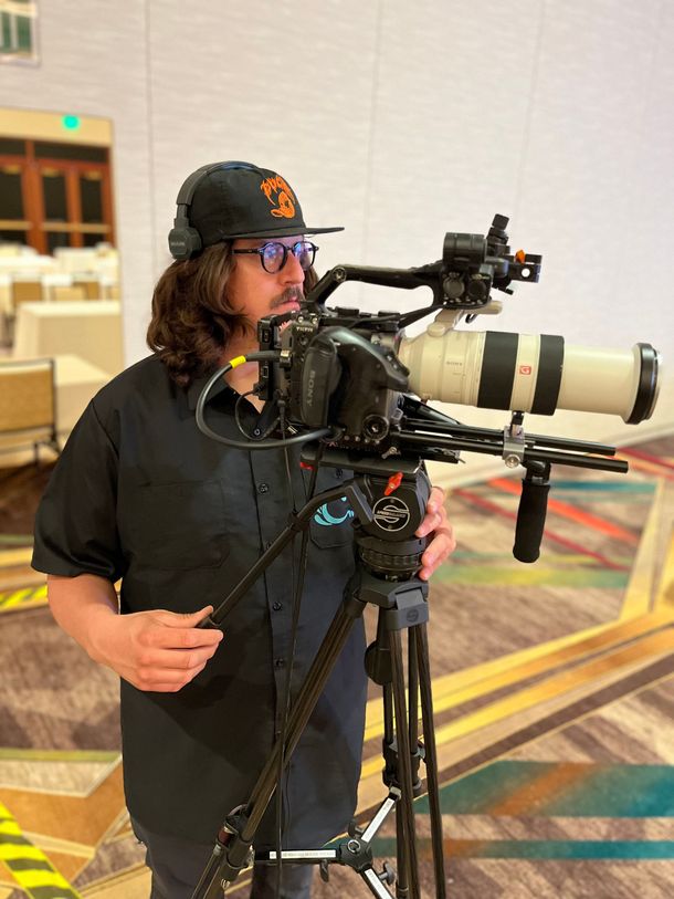 Camera operator beside a tripod-mounted telephoto lens in a conference hall
