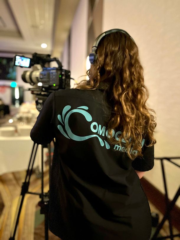 Camera operator filming in a hallway, wearing a black shirt with a teal logo.