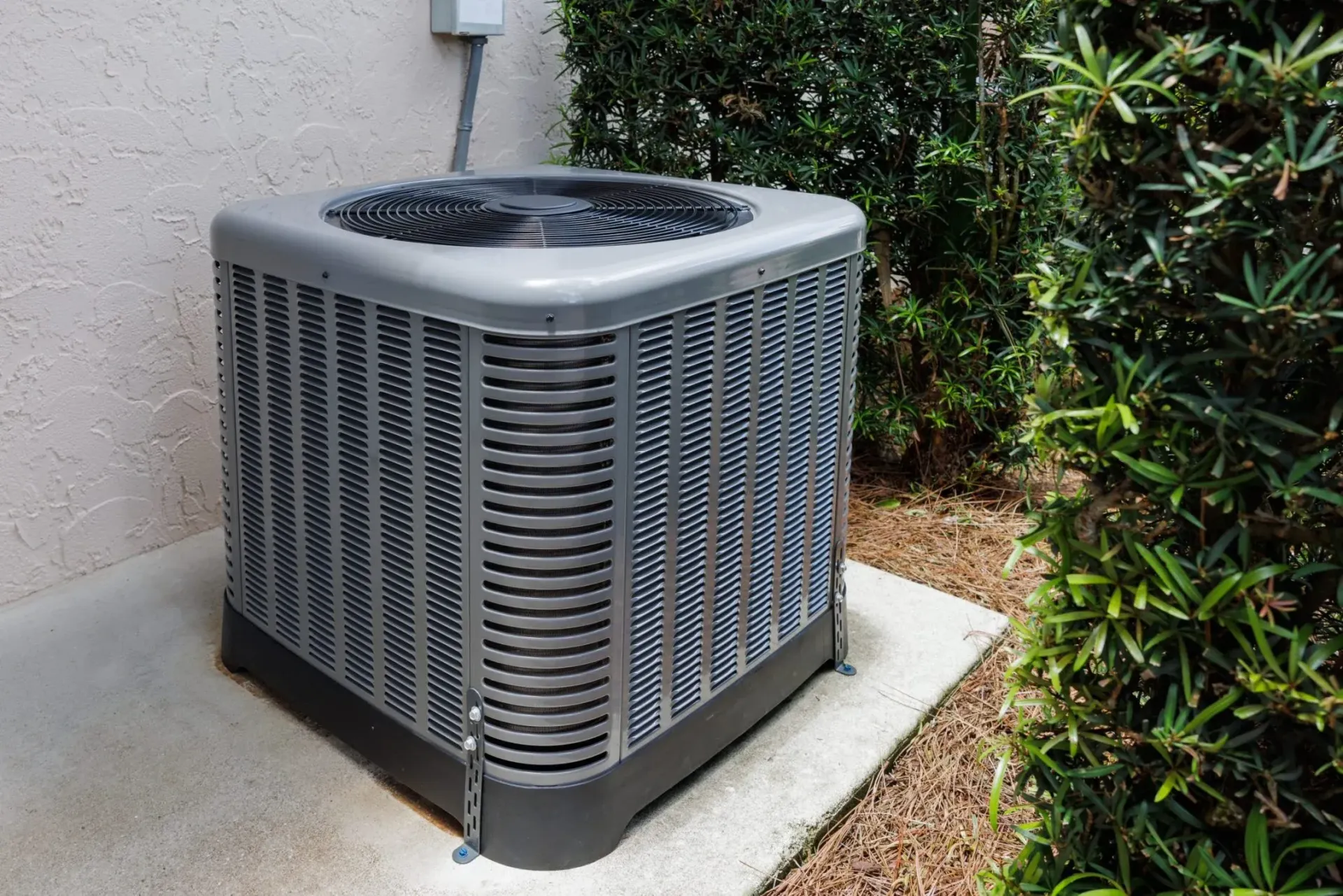 Outdoor air conditioner unit beside a house on a concrete pad, next to shrubs and a wall.