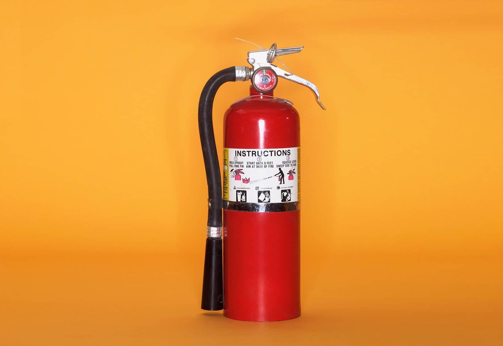 Extintor de incendios de color rojo sobre fondo naranja, con manguera negra adjunta y etiqueta de instrucciones.