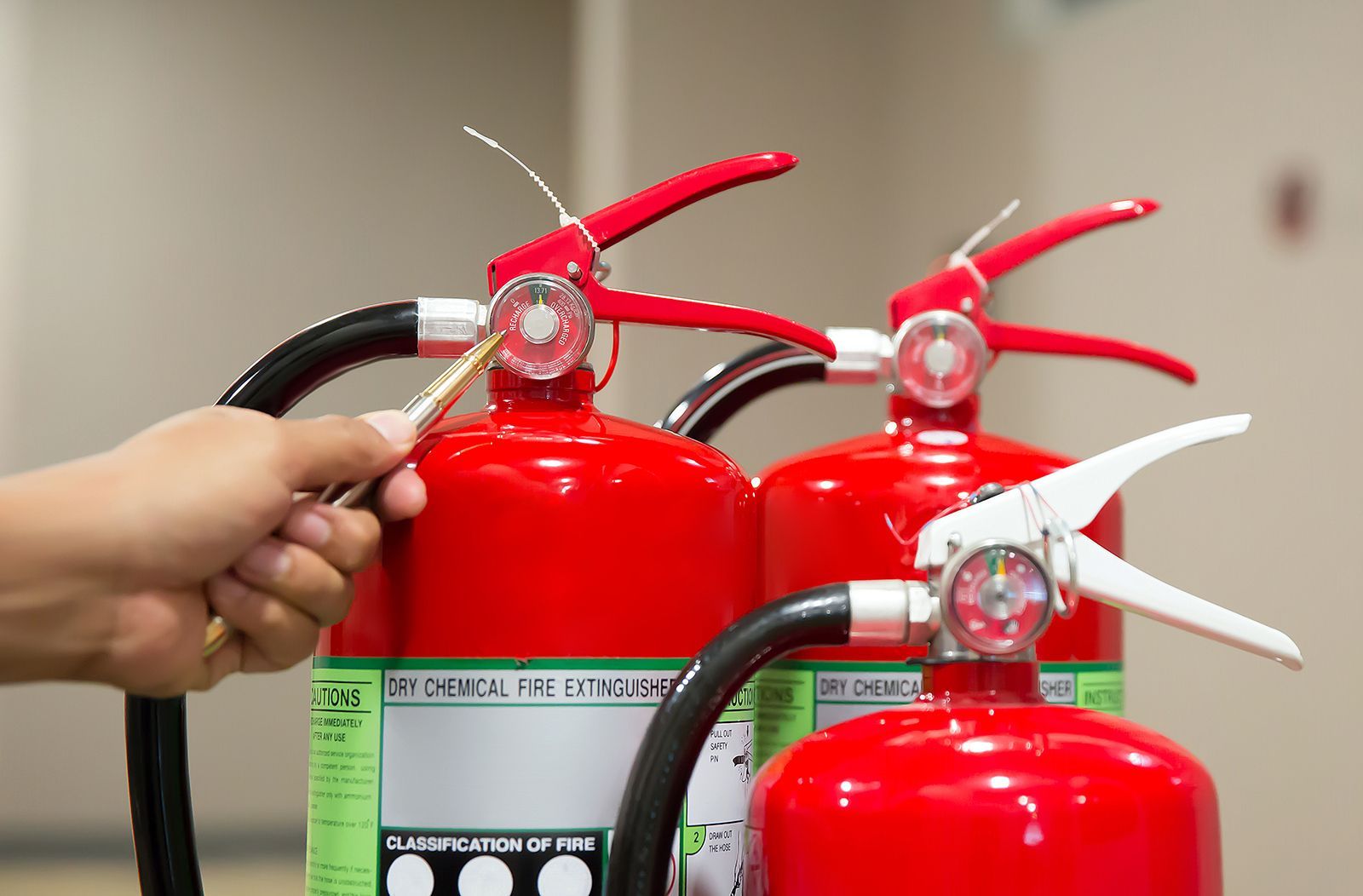 Una mano usa un alfiler para inspeccionar el sello de seguridad de un extintor. Tres extintores rojos están alineados.