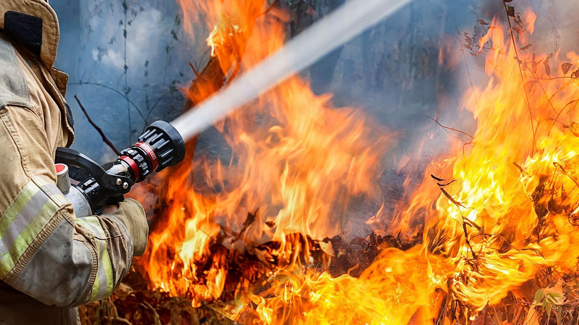 Bombero rociando agua sobre un gran incendio, las llamas envuelven una pared.
