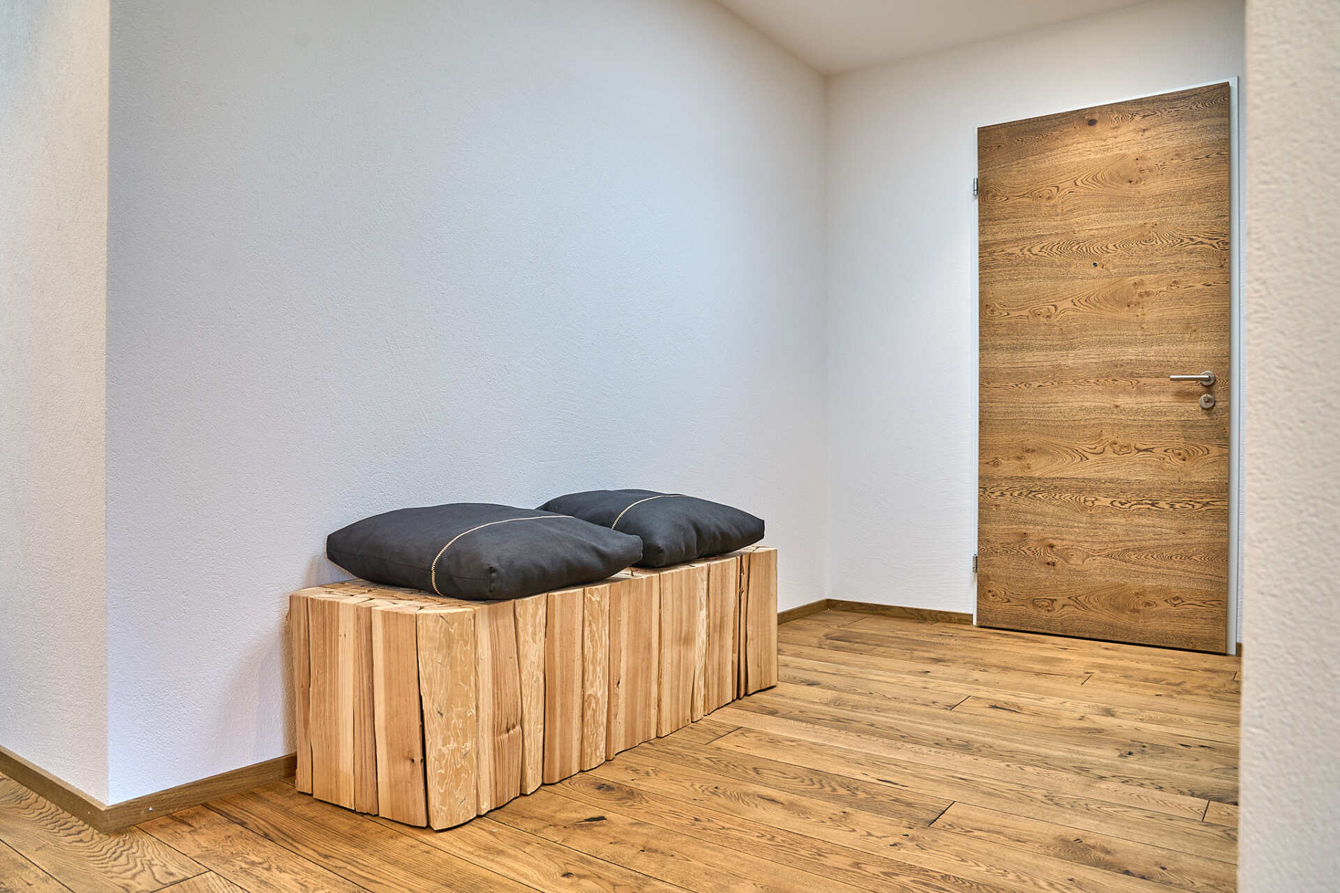 Eine Holzbank mit zwei Kissen darauf steht in einem Flur neben einer Tür.