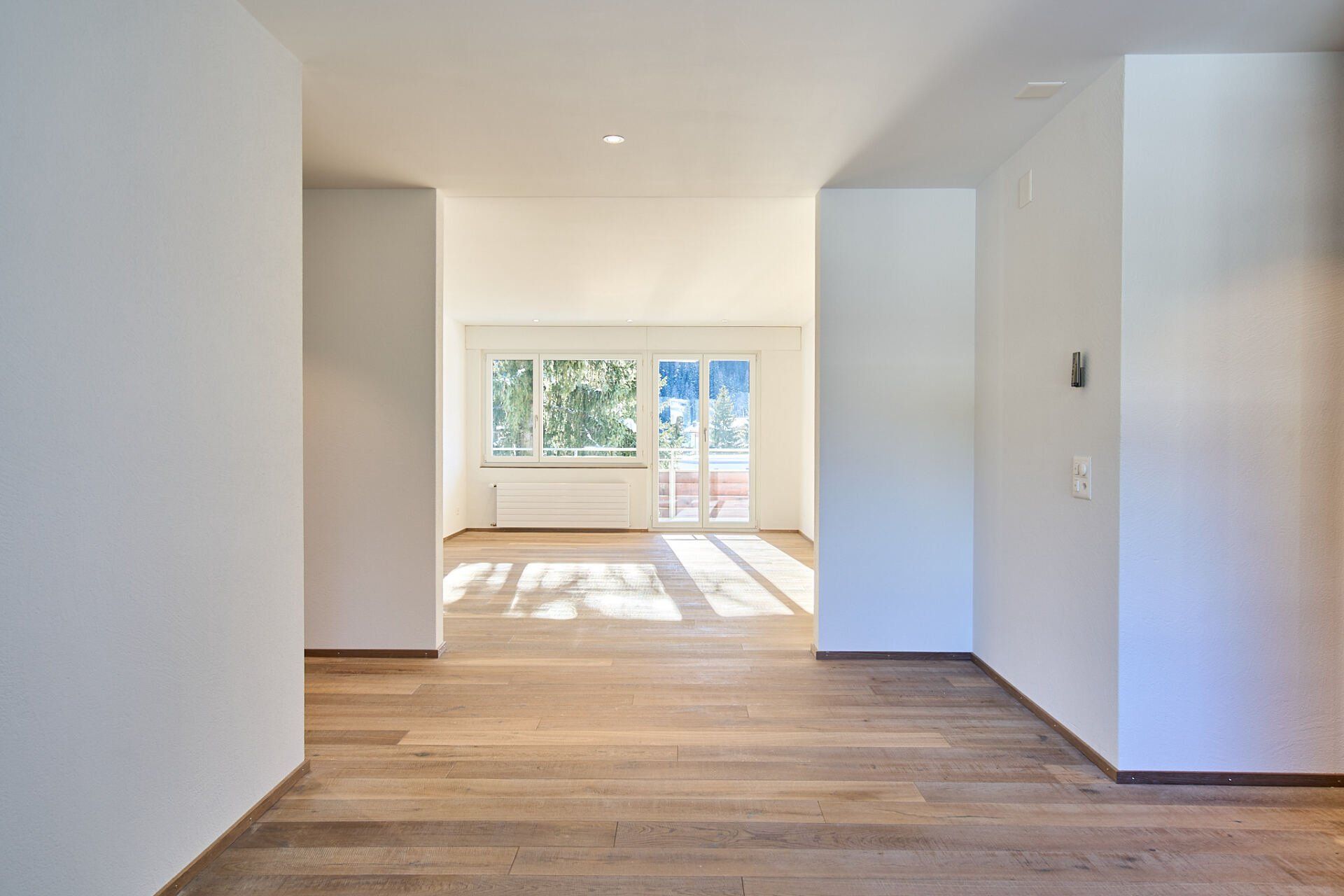 Ein leerer Flur mit Hartholzböden und weißen Wänden, der zu einem Wohnzimmer führt.