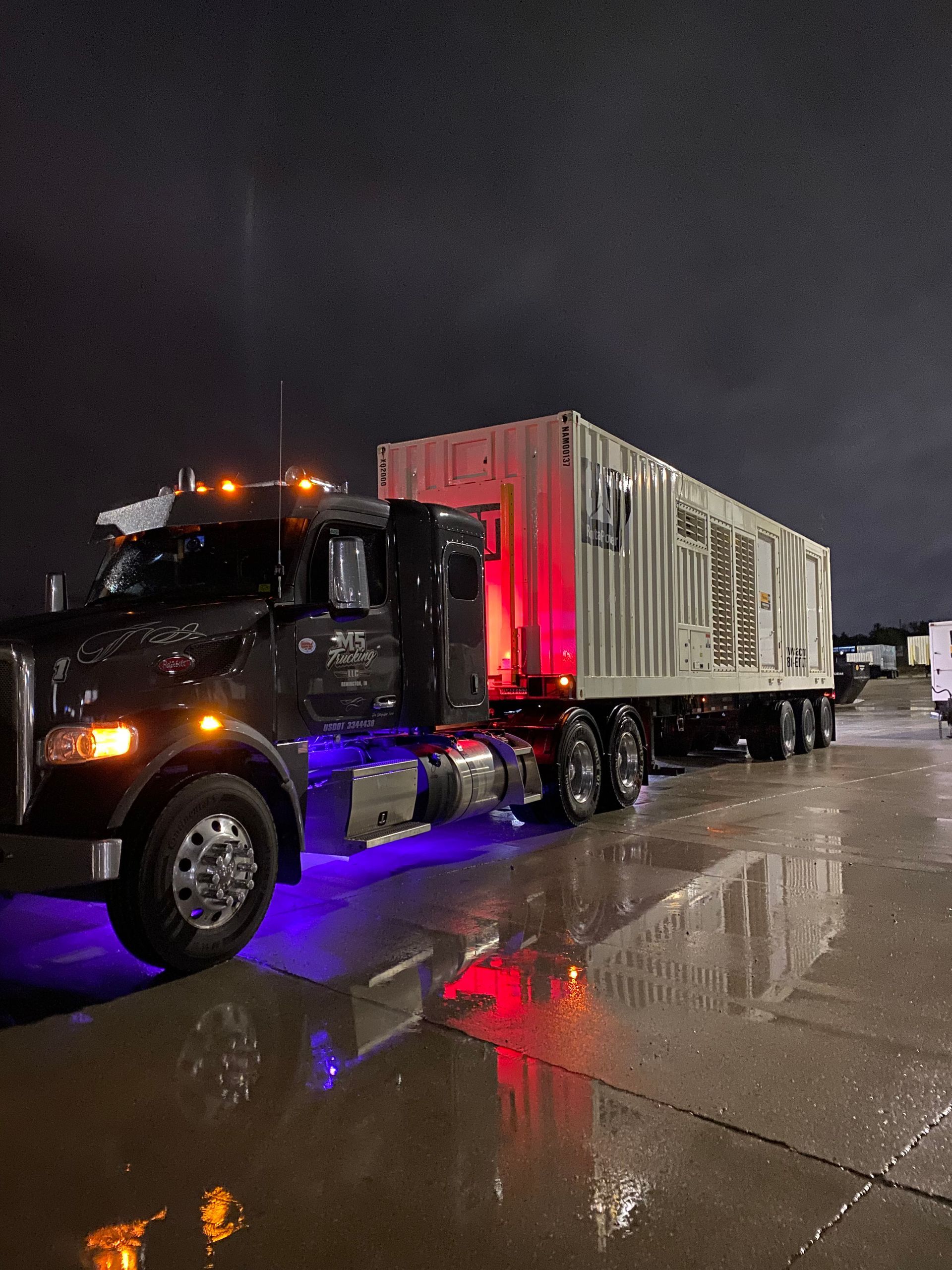 A semi truck with a trailer attached to it is parked in a parking lot at night.