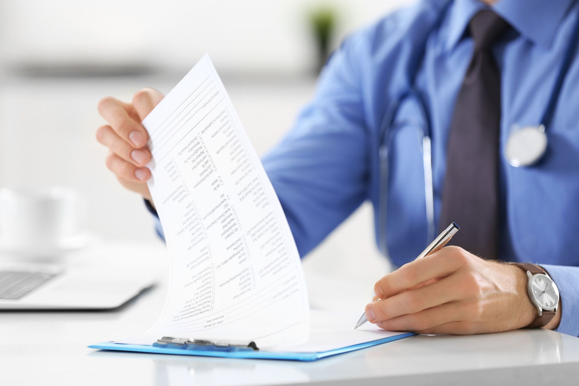 Doctor in blue shirt, writing on a medical form at a desk, stethoscope around neck.