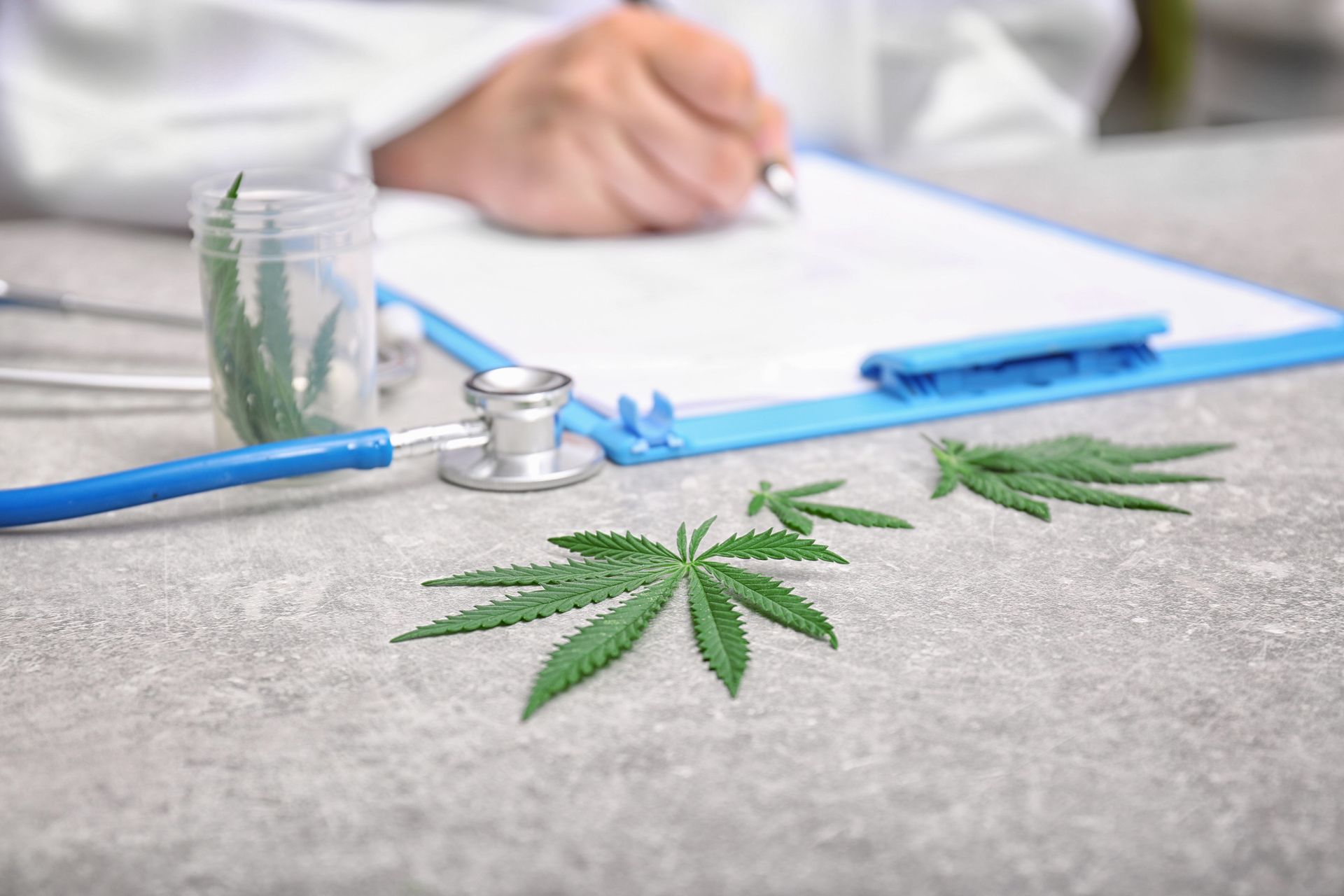 Doctor writing on clipboard with marijuana leaves and stethoscope on a table.