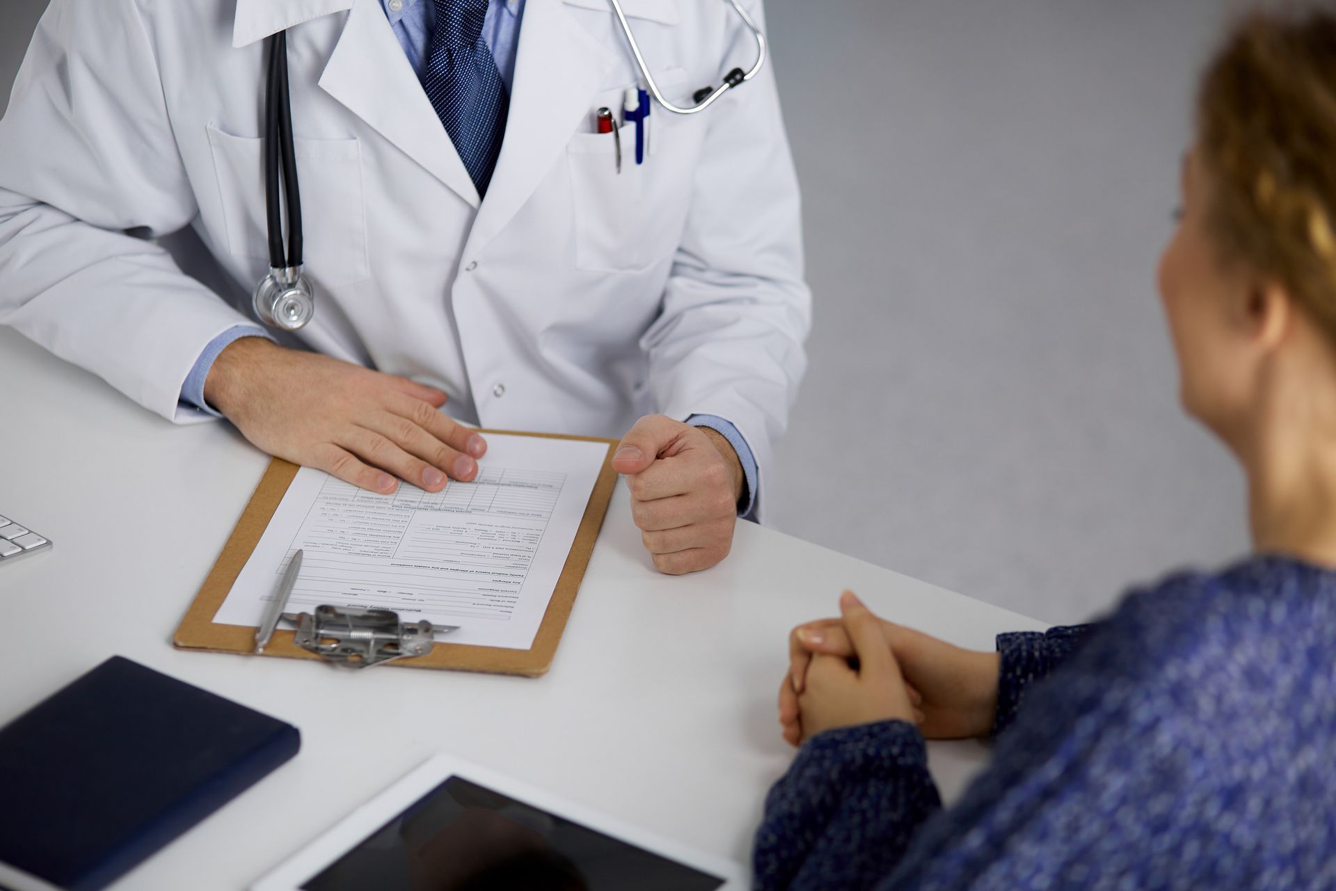 Doctor in white coat and patient at desk, doctor holding clipboard, listening.