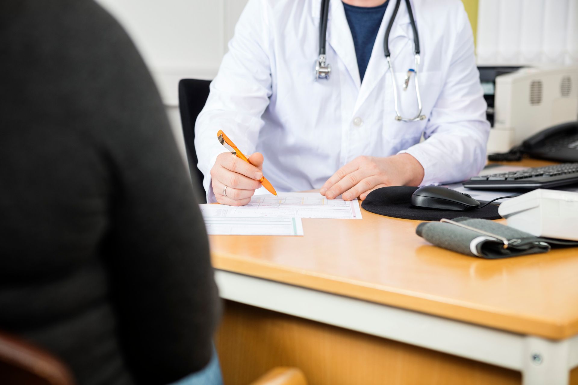 Doctor in white coat reviewing paperwork with a patient seated across a wooden desk.