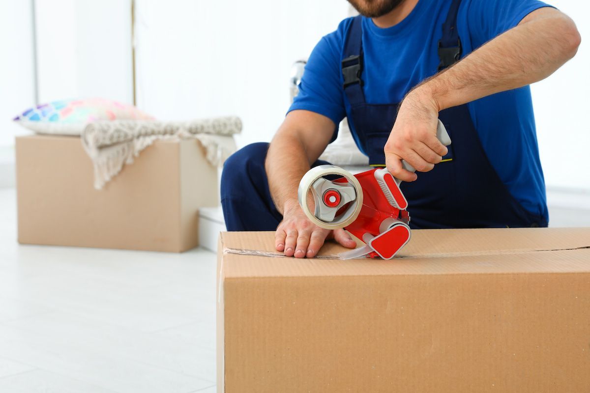 A Man Is Taping a Cardboard Box with Tape — Ocean Grove Removals In Colac, VIC