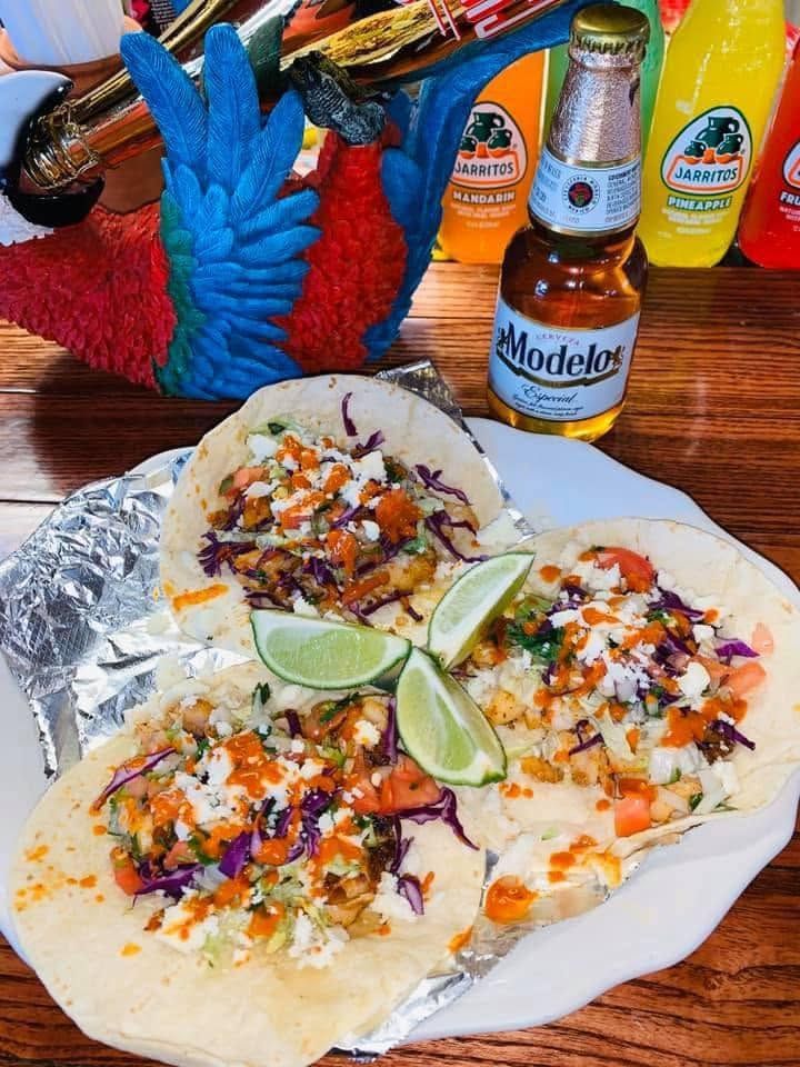 Mexican street tacos on foil with lime wedges, colorful napkins, and a Modelo beer bottle on a wooden table