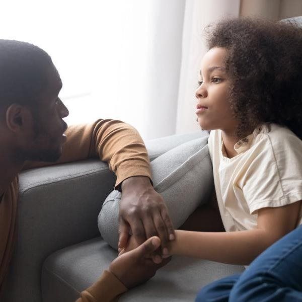 Man holding a child's hand, looking at her with concern, sitting on a couch near a window.