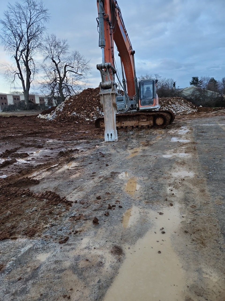 Excavator with jackhammer attachment working on muddy ground, overcast sky.