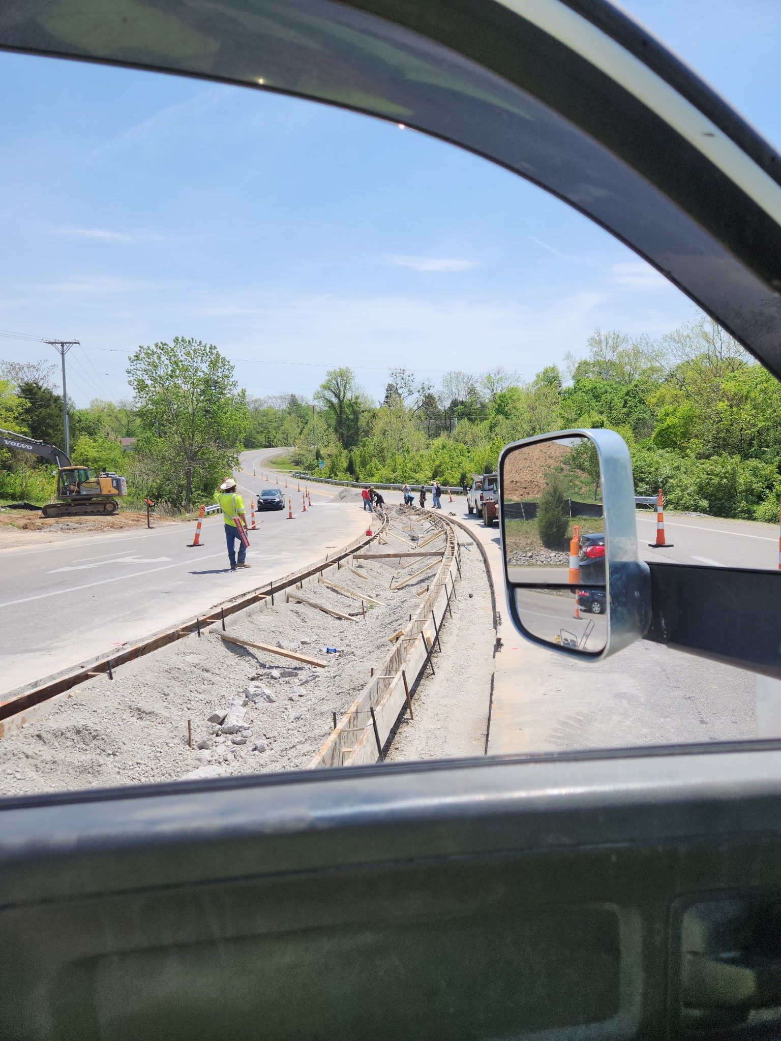 Road construction scene. A worker in a vest directs traffic. Gray roadbed being worked on under a bright blue sky.