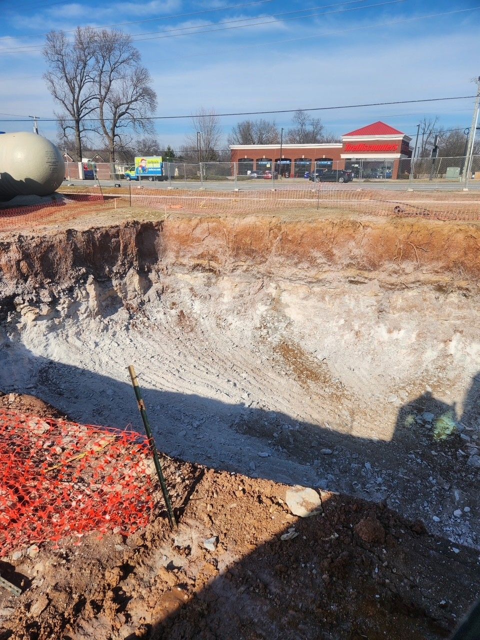 Construction site with a deep excavation, red and brown dirt. A building with a red roof is in the background.