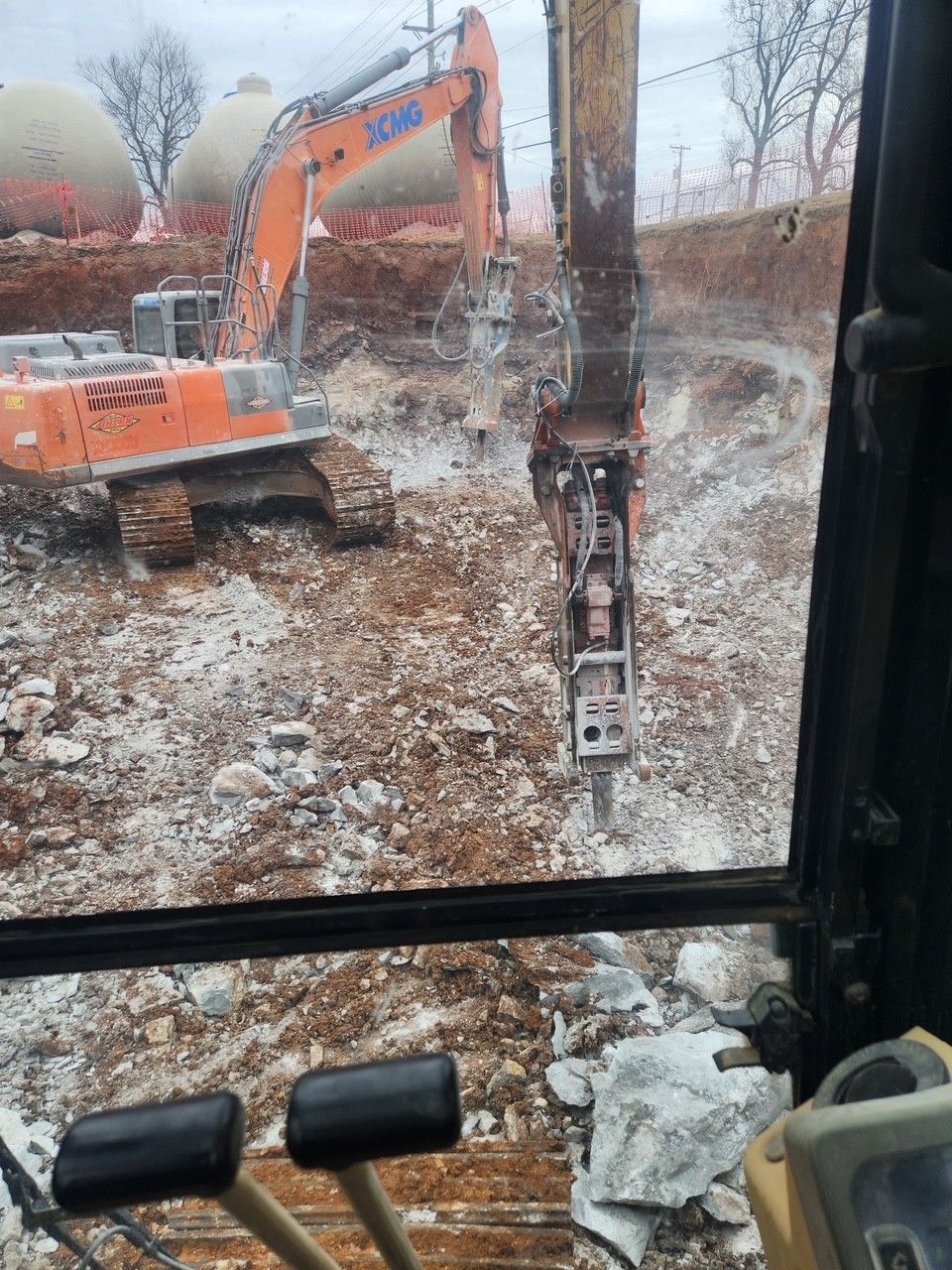 An excavator with a jackhammer attachment demolishes a rocky hillside; view from inside the cab.