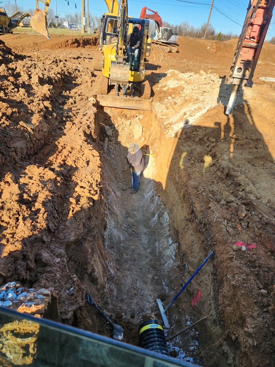 Construction site: Excavator digging a trench, worker standing inside. Earth and pipes are visible.