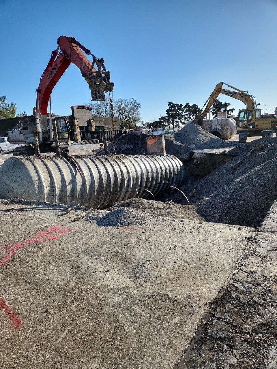 Construction site with large corrugated metal pipe, two excavators, and piles of gravel.