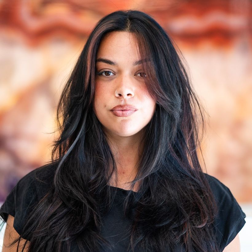 Donna con capelli lunghi e scuri, taglio scalato, che indossa un top nero. Sfondo sfocato.