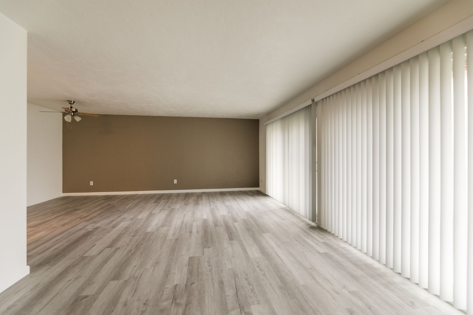 An empty living room with hardwood floors , blinds and a ceiling fan.
