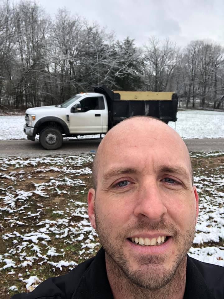 A man is smiling in front of a dump truck in the snow.
