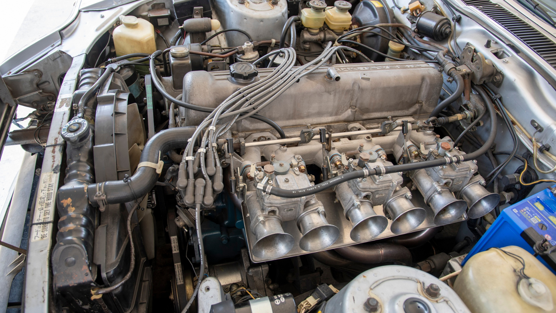 An open car hood reveals a Nissan L-series inline-six engine equipped with triple side-draft carburetors and velocity stacks.