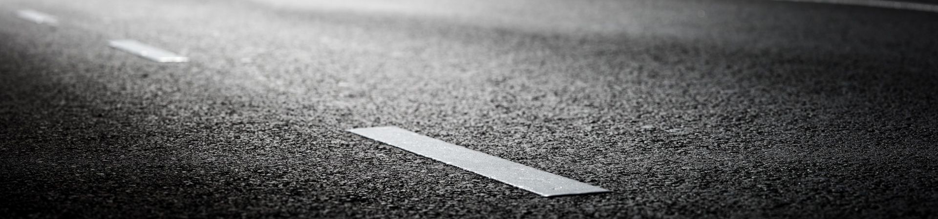 Un primo piano in bianco e nero di una strada con la segnaletica orizzontale tratteggiata bianca che si perde in lontananza.