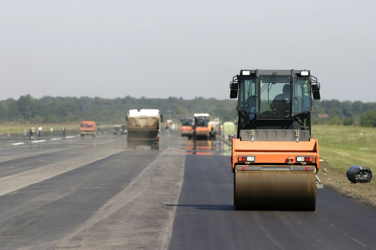 Un rullo compressore arancione è al lavoro su una pista aeroportuale in asfalto scuro, con veicoli da cantiere sullo sfondo.