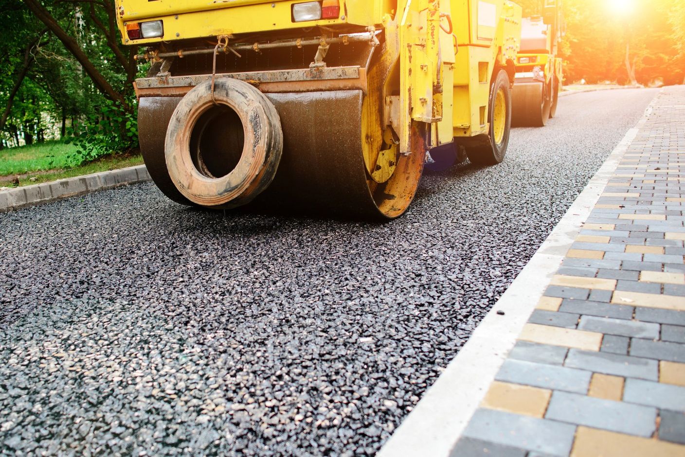 Un rullo compressore giallo passa sopra della ghiaia fresca su una strada accanto a un marciapiede asfaltato al tramonto.