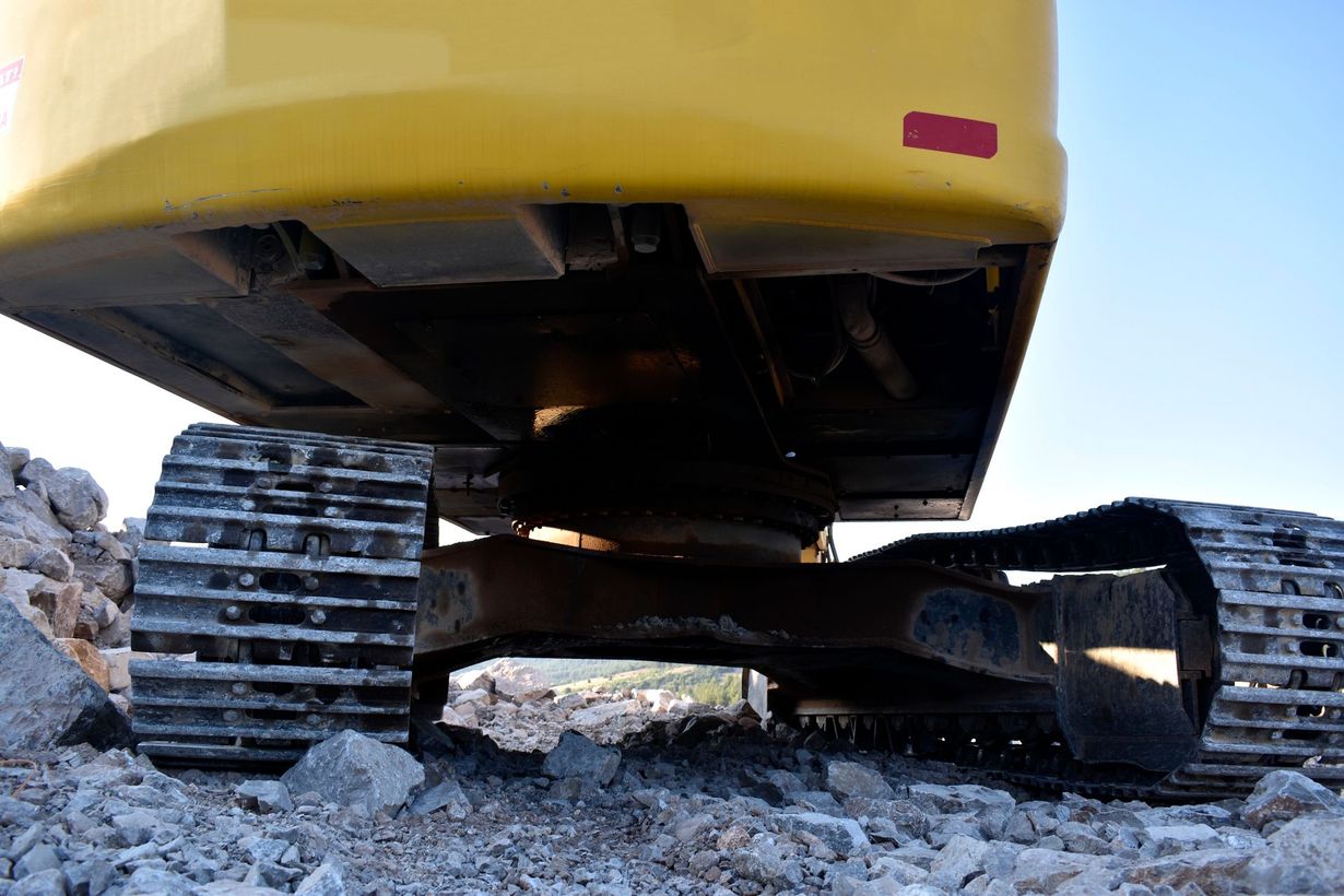 Una ripresa dal basso che mostra i cingoli e il sottocarro di un escavatore giallo parcheggiato su un terreno roccioso e irregolare.