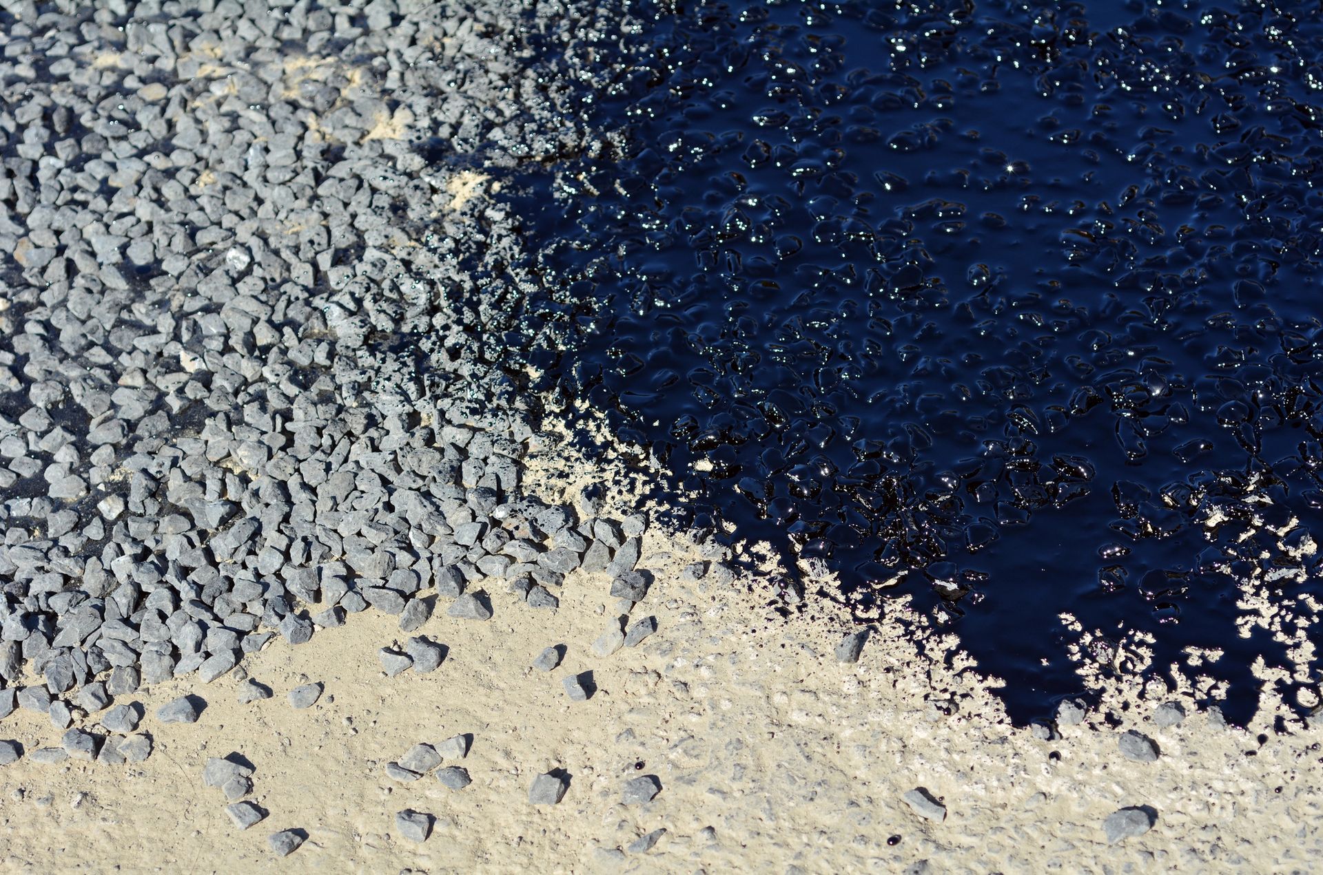 Una vista dall'alto che mostra la transizione da ghiaia grigia e sciolta sulla sinistra a una zona di asfalto scuro e lucido sulla destra.