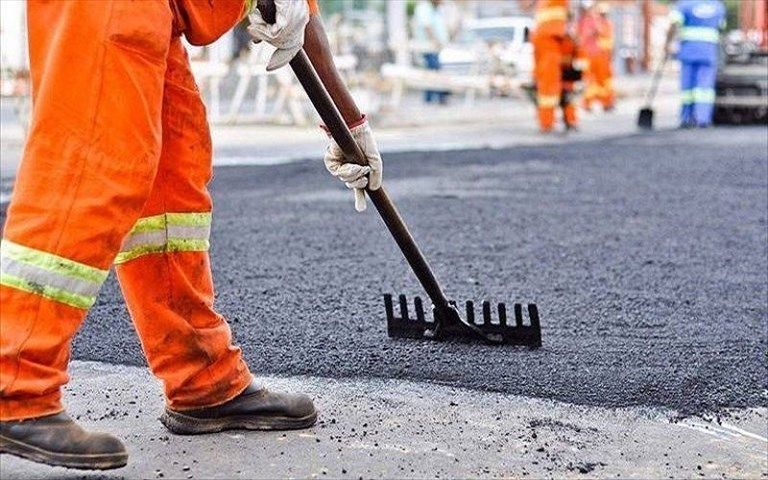 Un operaio con indosso una tuta arancione ad alta visibilità utilizza un rastrello per livellare l'asfalto fresco in un cantiere stradale.