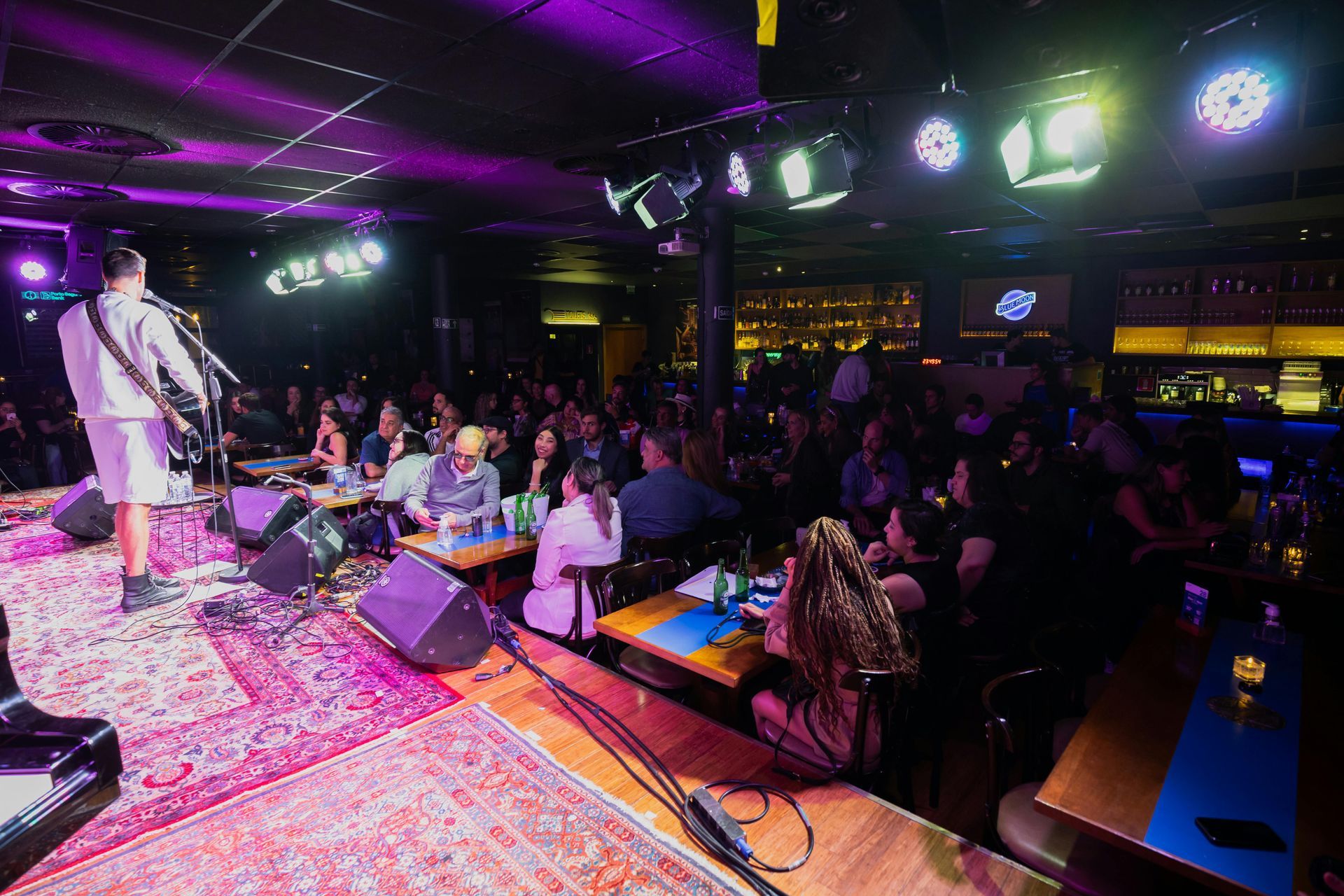 Live music venue: band on stage, audience seated at tables, purple and blue lighting.