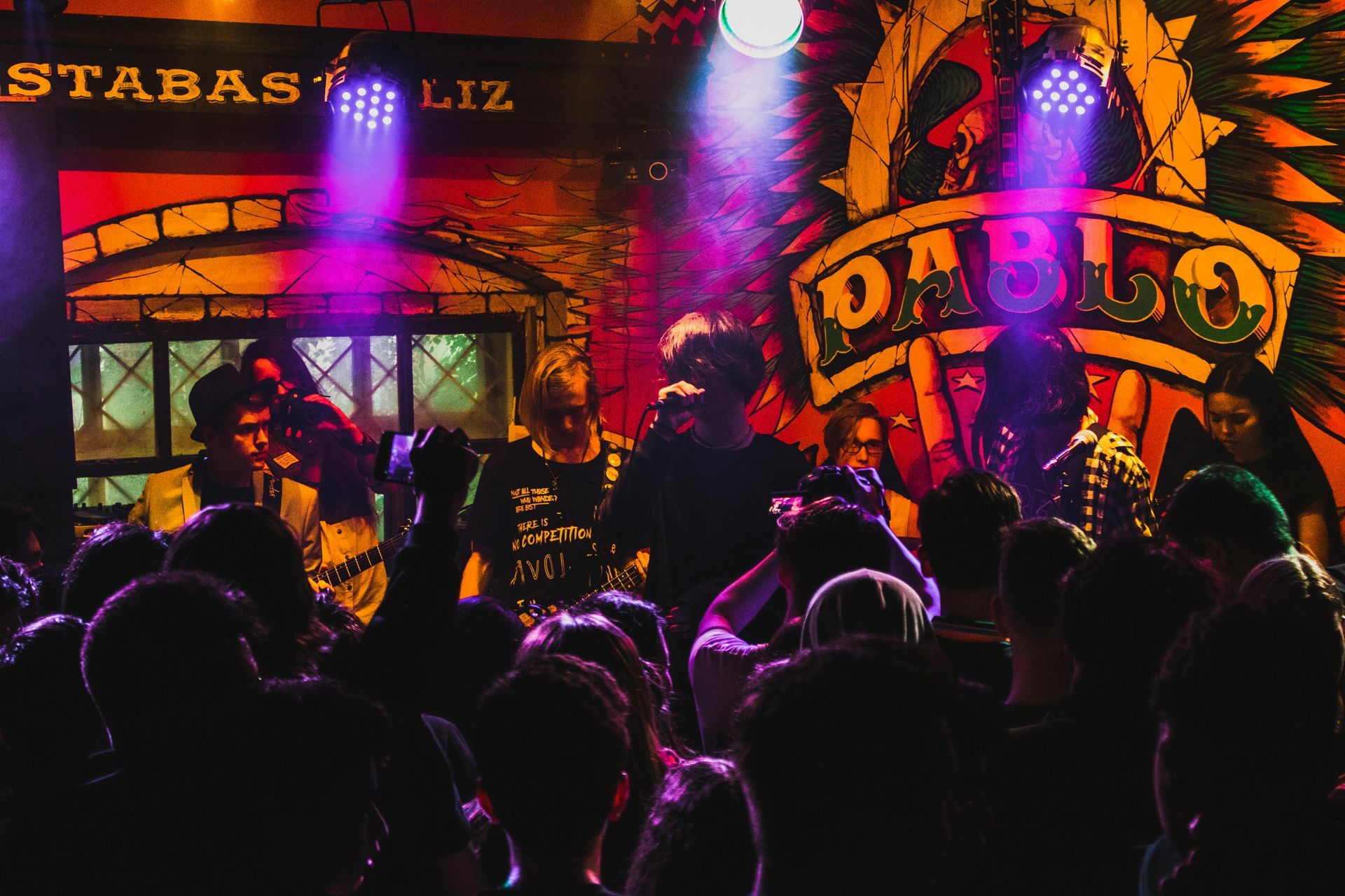 Band performs on stage in a crowded venue lit by spotlights, with 