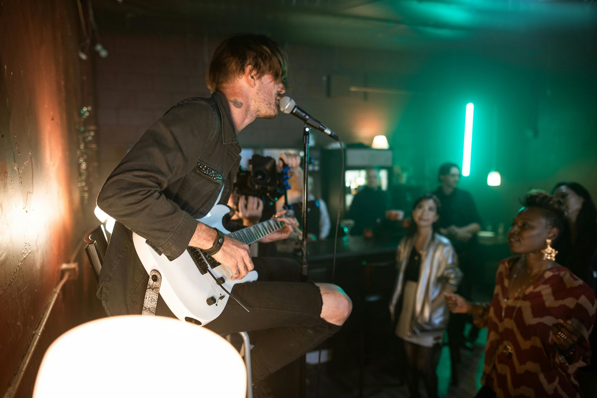 Man singing and playing electric guitar on stage at a venue. Green and orange lighting, audience visible.