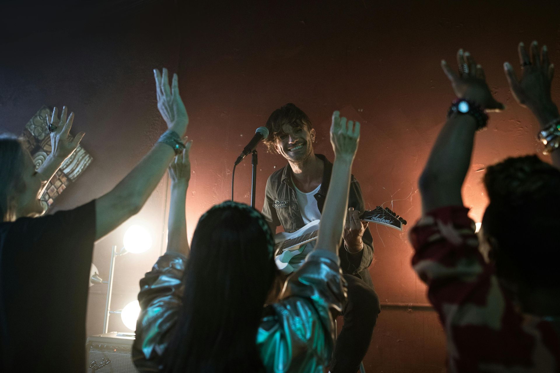 Band performing on stage, guitarist singing with arms raised, audience with hands up, stage lights.