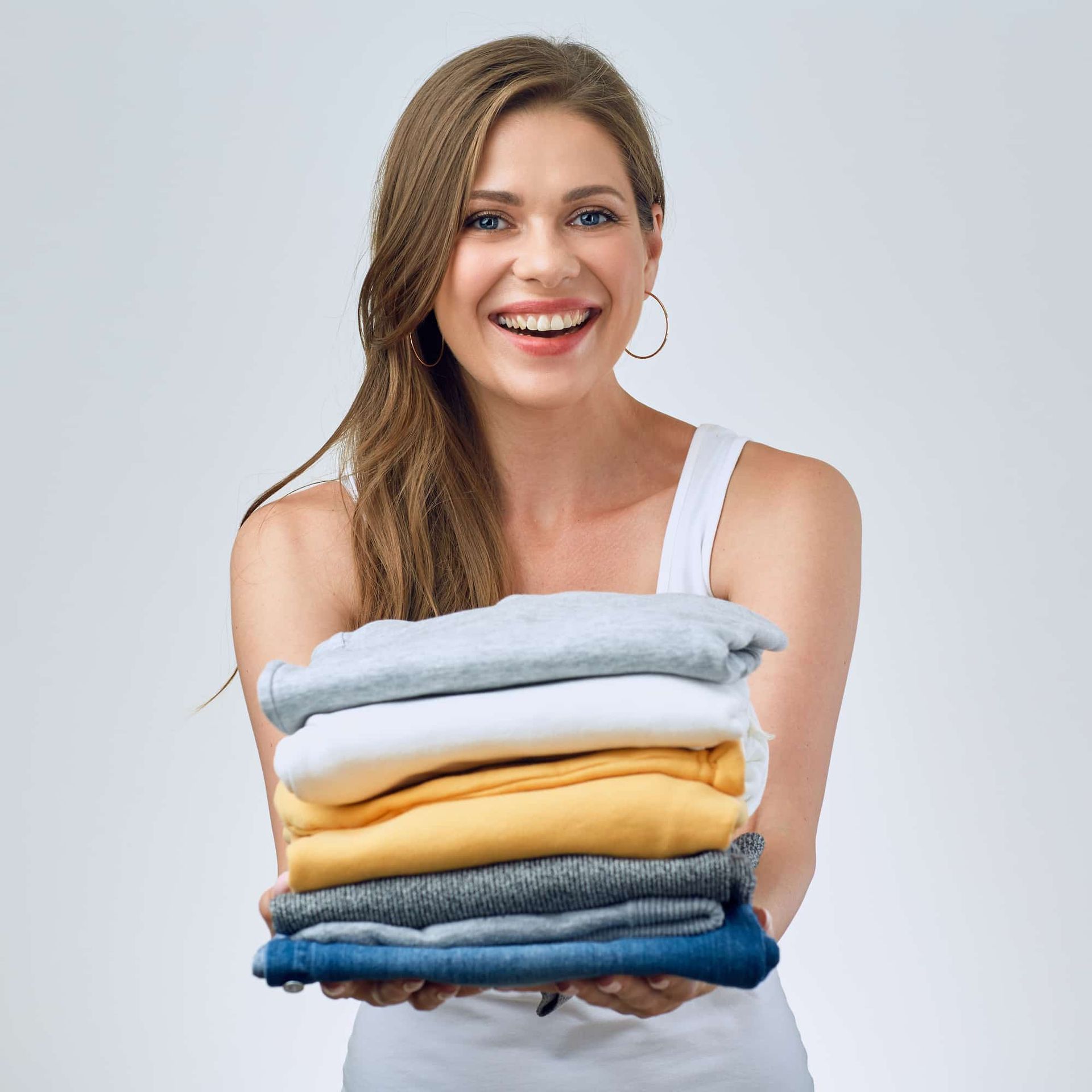 Woman smiling, holding a stack of neatly folded clothes; gray background.