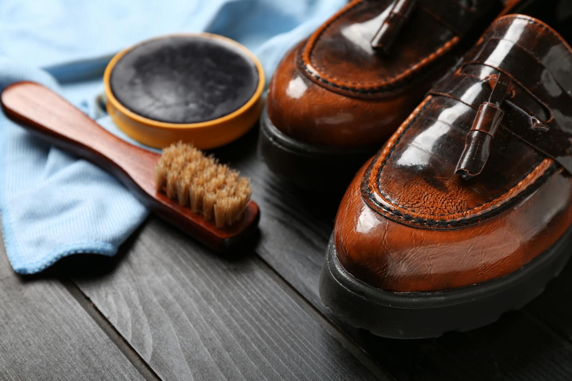 Brown leather shoes with shoe polish, brush, and cloth on a dark wooden surface.